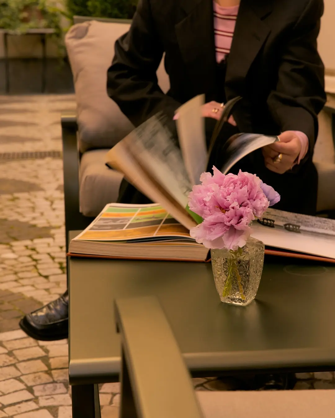 Someone reading at an outdoor table beside a glass of peonies