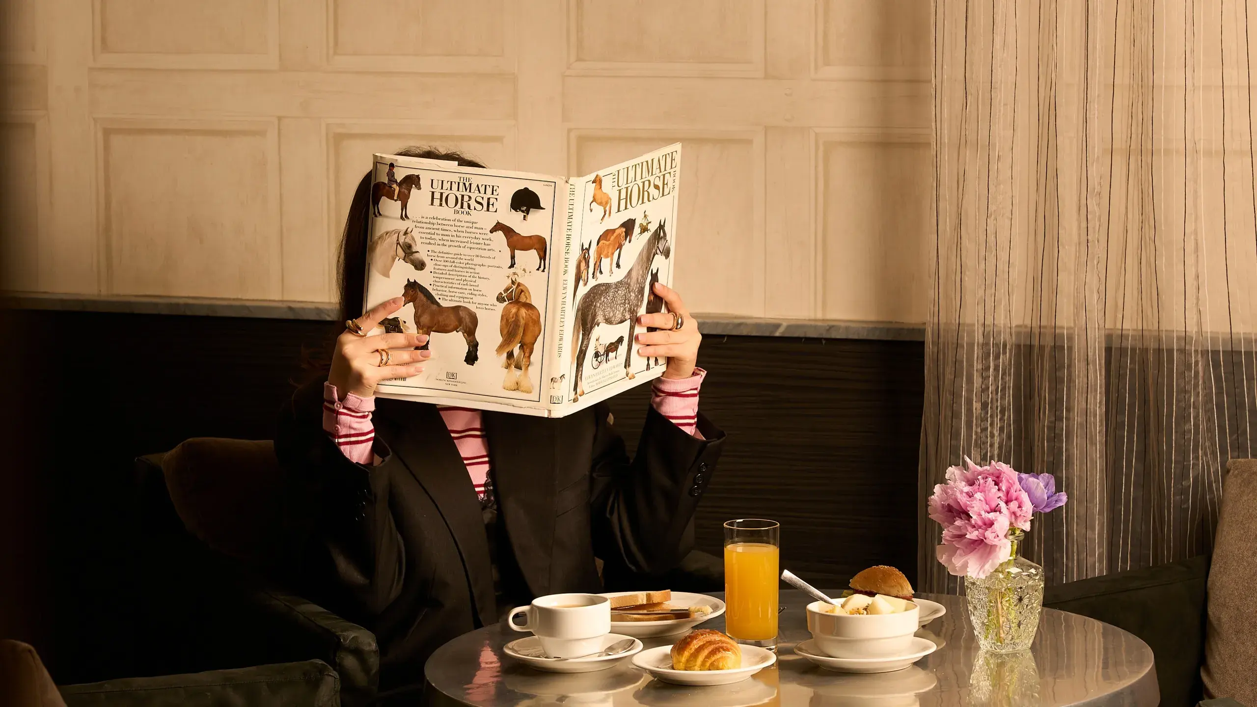 A girl at breakfast holding a book about horses in front of her face