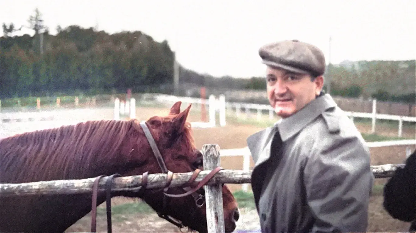 An archival photograph of a man in a flat cap standing beside a horse at a rail
